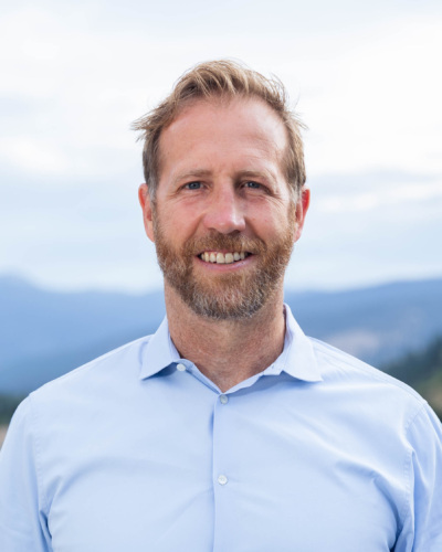 Headshot of Jake Harriman, a white man with a salt and pepper beard standing in front of scenic mountains