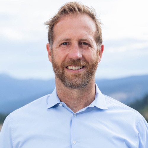 Headshot of Jake Harriman, a white man with a salt and pepper beard standing in front of scenic mountains