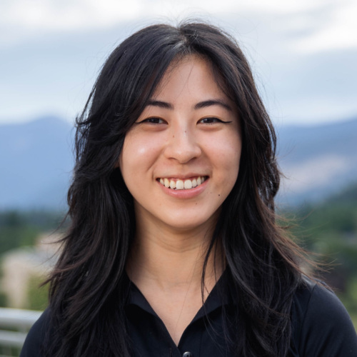 Headshot of Maina Chen, an Asian woman with long dark wavy hair in a black dress shirt with mountains in the background.