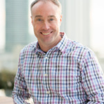 Headshot of a white, middle-aged man smiling in a checkered button up shirt