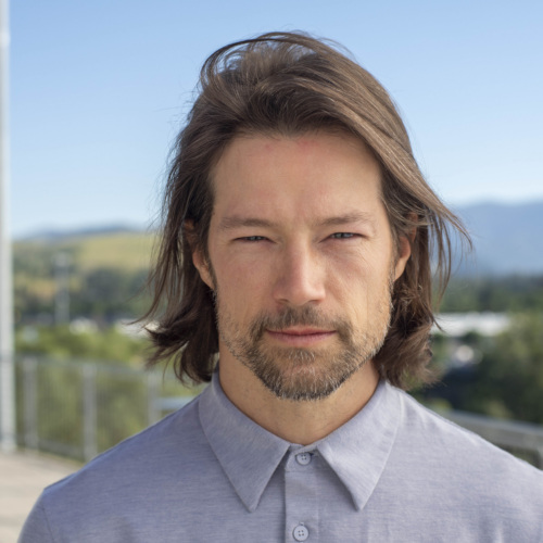 Headshot of Trip O'Connell, a Caucasian man with windswept light brown hair in a blue dress shirt with mountains in the background.