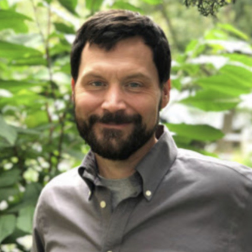 Picture of Alex Ireland, a white middle aged man with brown hair and a beard standing in front of trees.